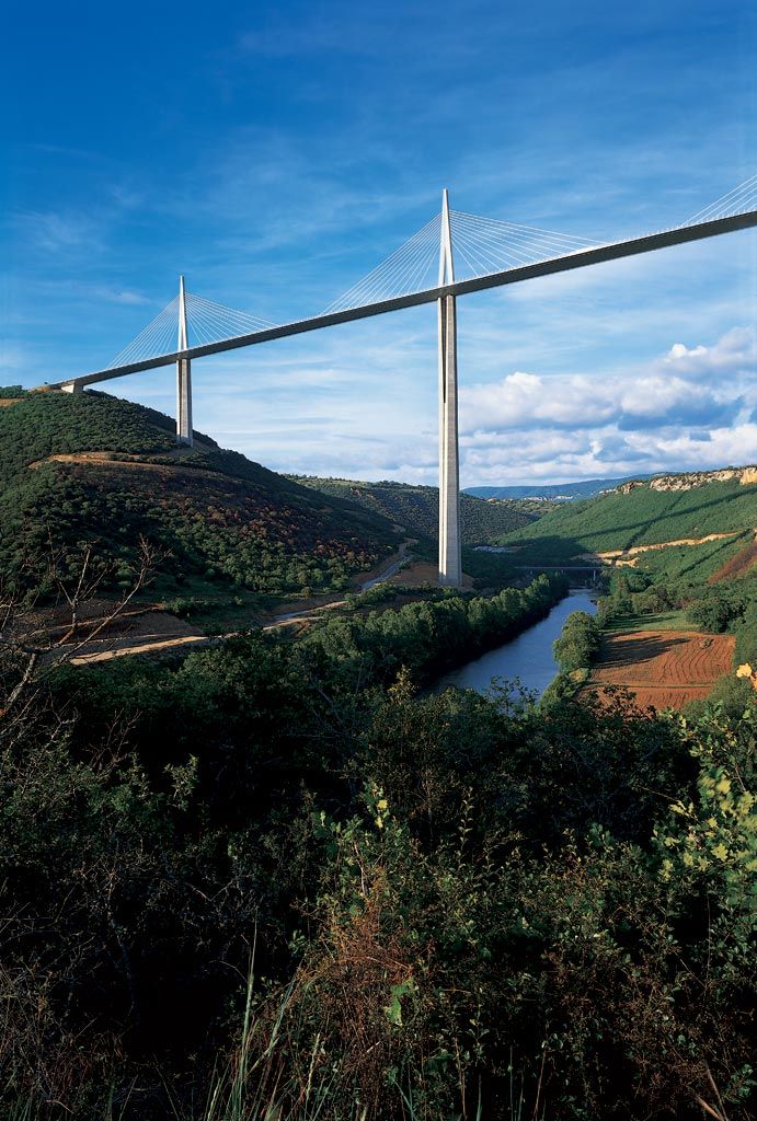 Tall cable-stayed bridge over a lush green valley and winding river under a clear blue sky.