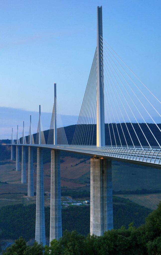 Modern cable-stayed bridge spanning a wide valley, with tall pylons against a clear blue sky.