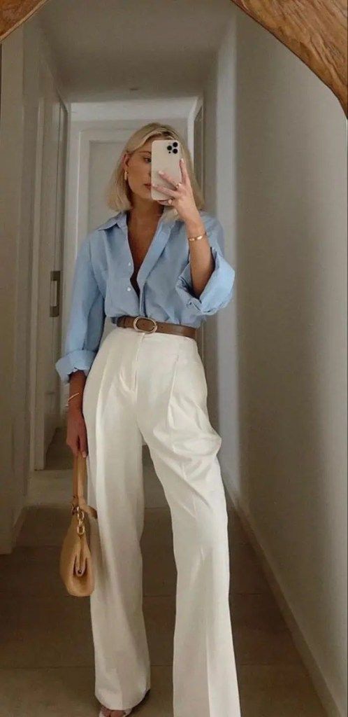 Fashionable outfit: woman in blue shirt, white pants, holding phone and beige handbag, standing in hallway mirror selfie.