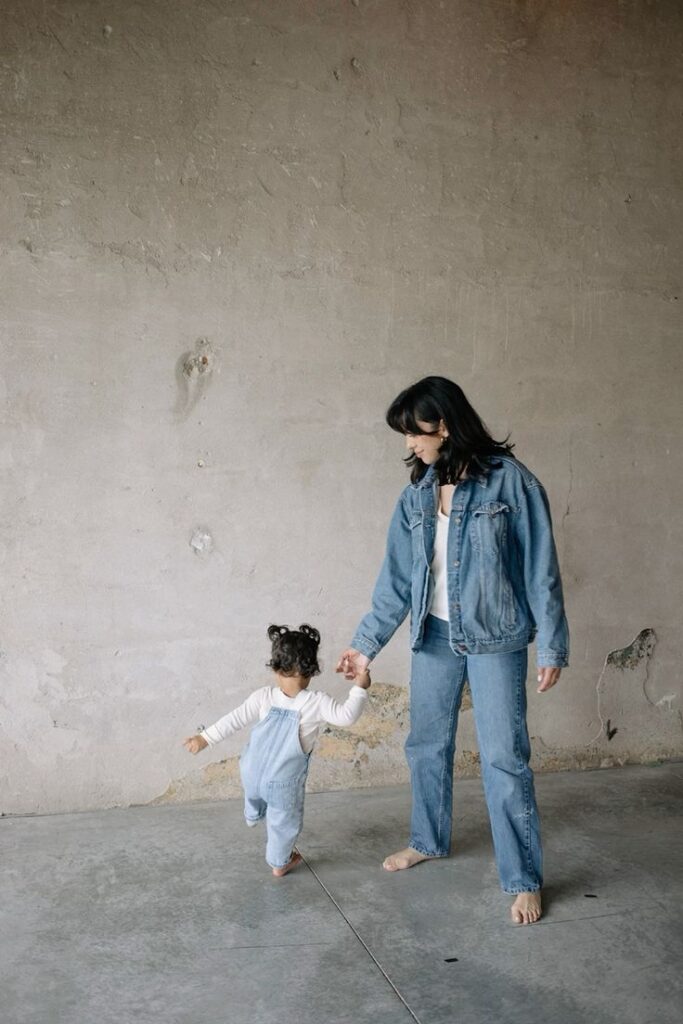 Mother and child in denim hold hands, walking indoors on a concrete floor with a textured wall background.