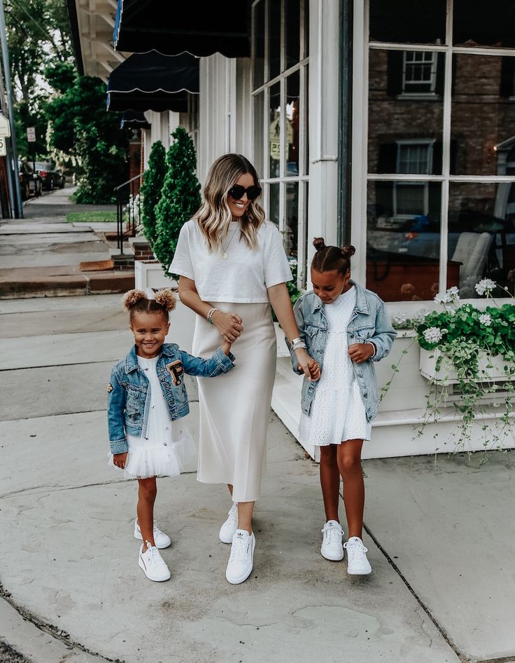 Mother and two daughters in stylish white outfits and denim jackets walking happily on a city street.