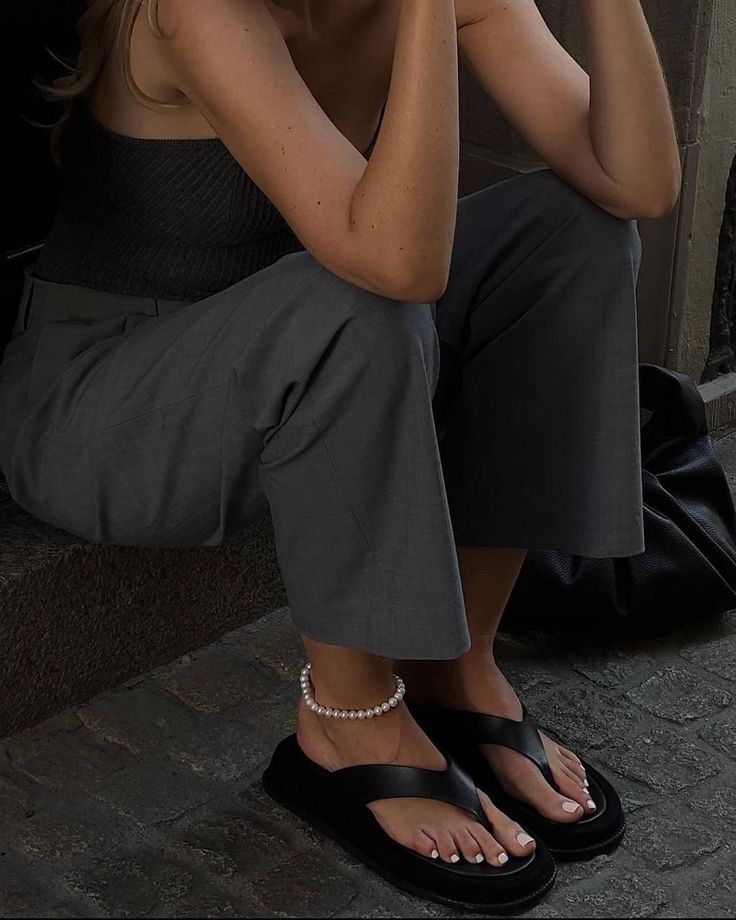 Woman sitting in gray pants and black sandals, wearing a pearl anklet, with a black bag on cobblestone pavement.