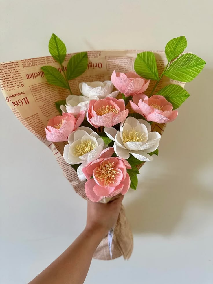 Bouquet of pink and white paper flowers with green leaves wrapped in newspaper-style paper.