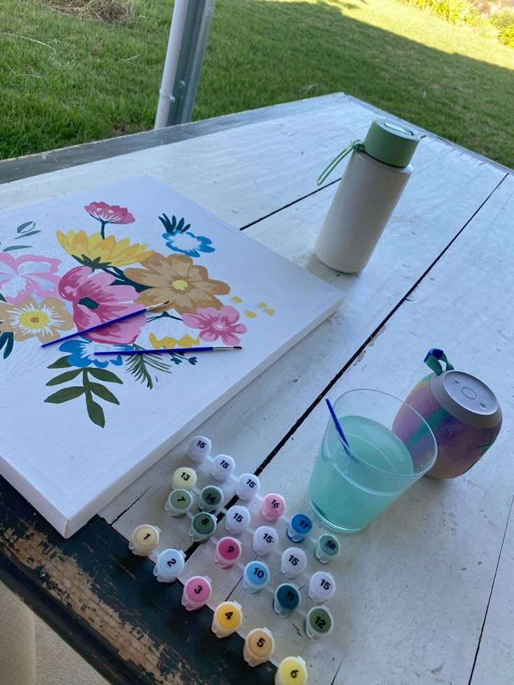 Paint-by-numbers kit with floral artwork on a wooden table, outdoors with a drink and speaker nearby.