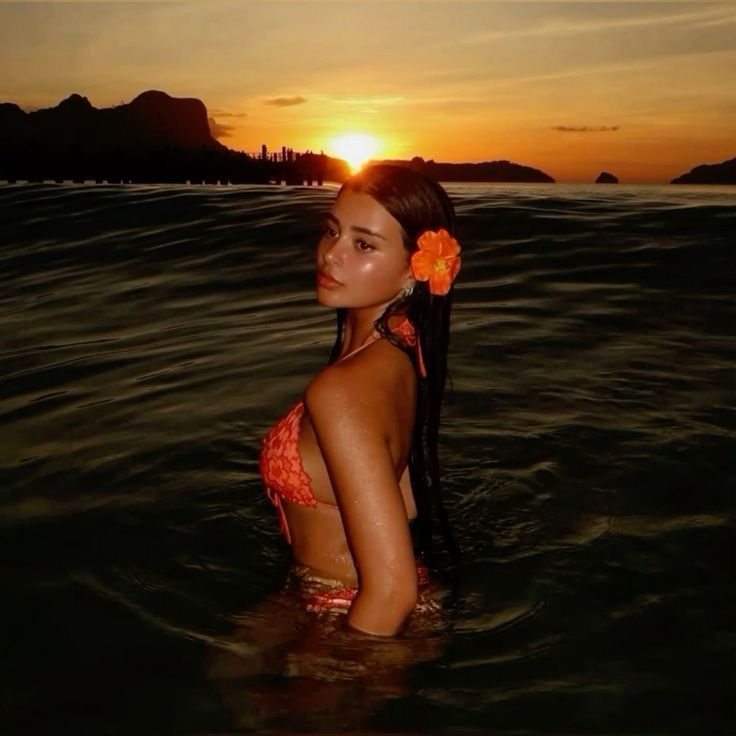 Woman in orange bikini with orange flower in hair at sunset, standing in sea, scenic view.