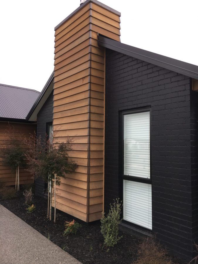 Modern house with black brick and wood siding, featuring a prominent chimney and surrounding landscaping.