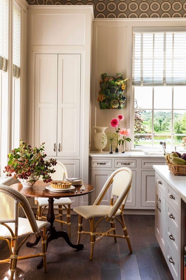 Cozy kitchen nook with wicker chairs, round wooden table, and white cabinets, overlooking a green landscape through a large window.