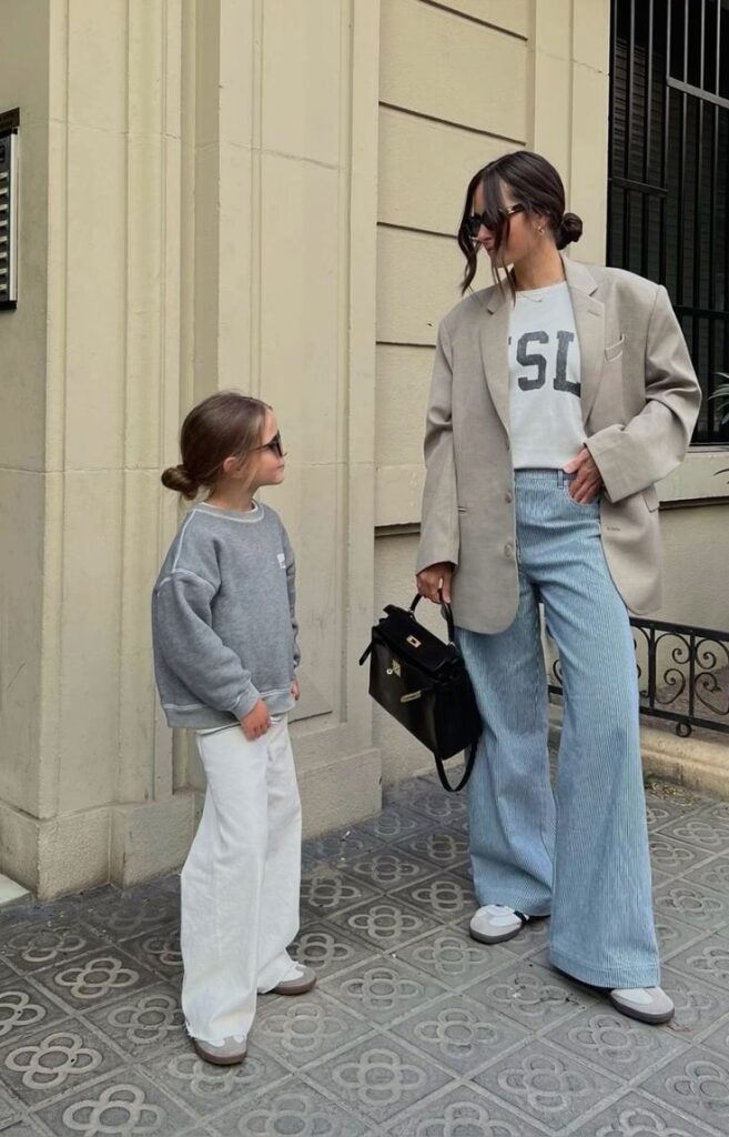 Mother and daughter in trendy outfits, wearing sunglasses, standing on a patterned sidewalk, smiling at each other.