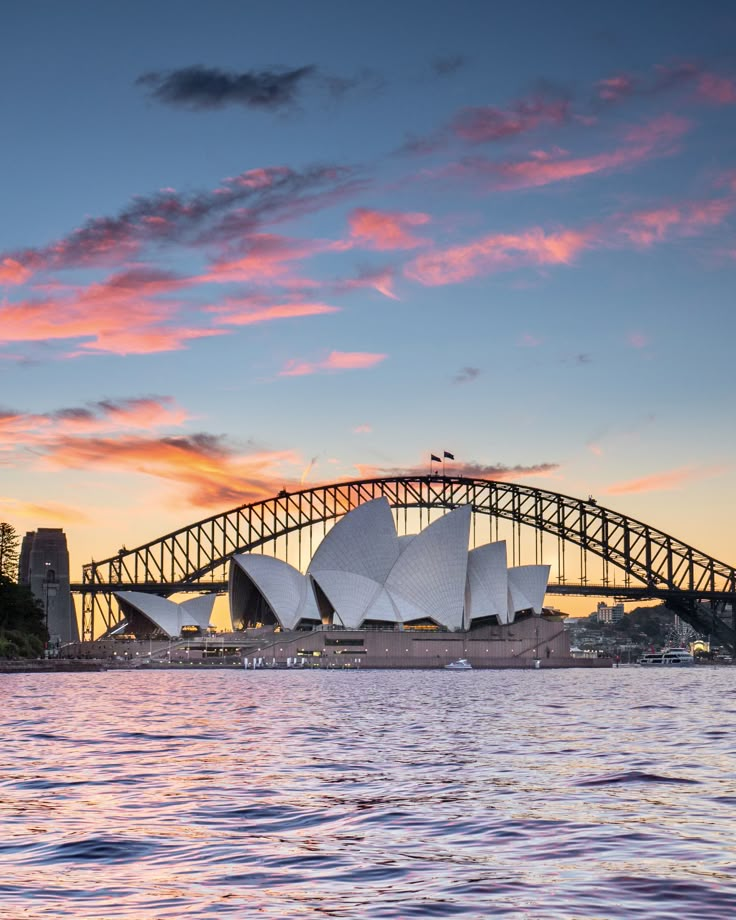 Sydney Opera House and Harbour Bridge at sunset, reflecting on the water, with vibrant pink and orange clouds.