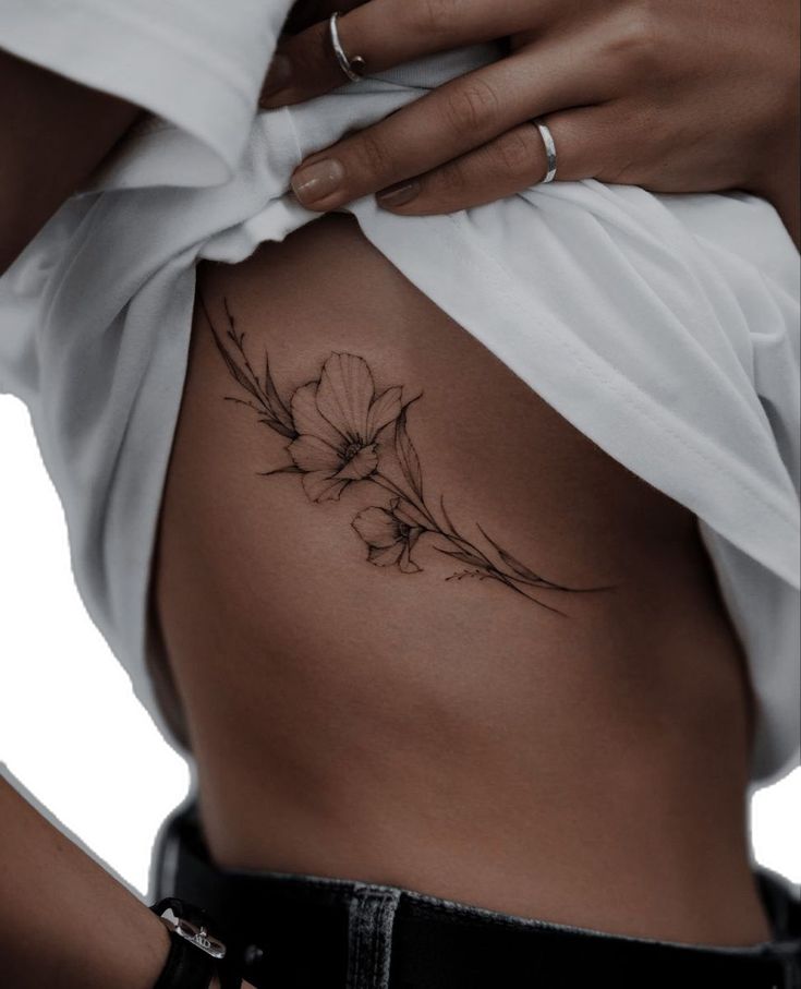 Floral side tattoo on woman's ribcage, white shirt lifted, showcasing delicate line art design.