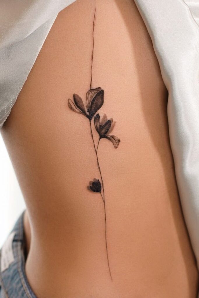 Minimalist floral tattoo on a person's side, featuring delicate black line art with blossoming flowers.