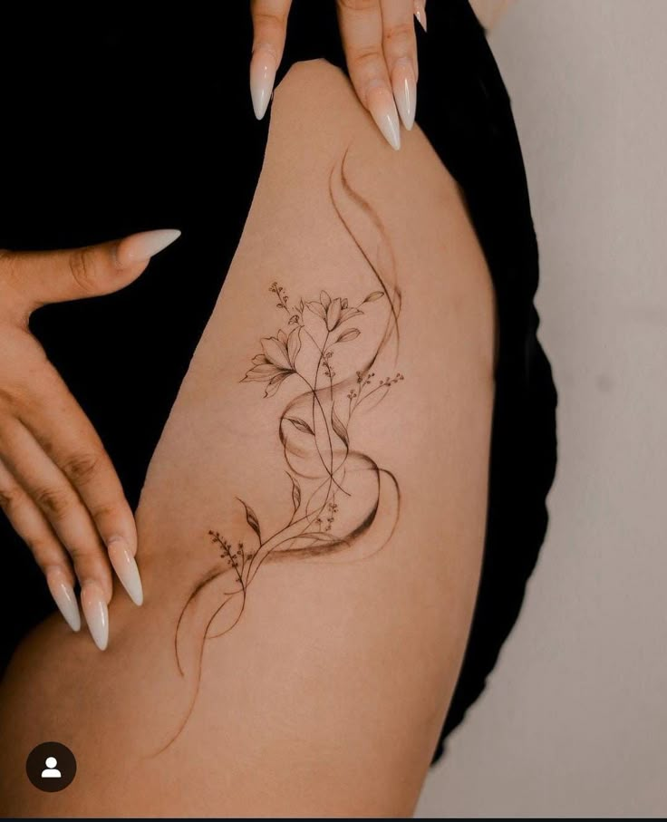 Delicate floral tattoo on thigh with intricate leaves and blossoms, accented by long fingernails and black attire.