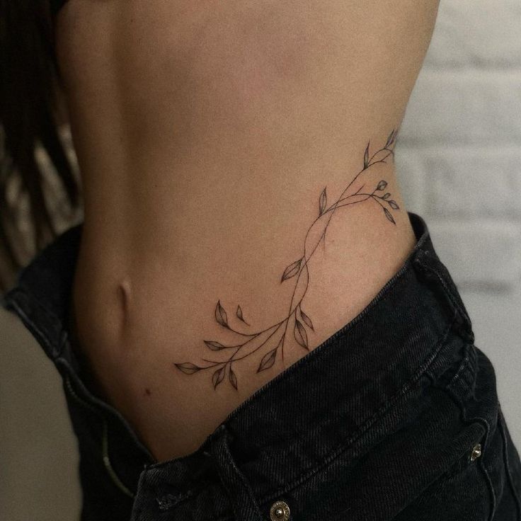 Minimalist vine tattoo on a woman's side torso, styled with dark jeans.