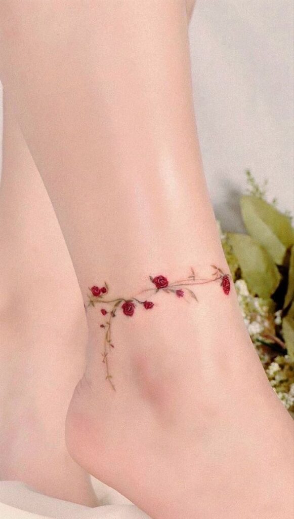 Minimalist ankle tattoo with delicate red roses and green vines on pale skin.