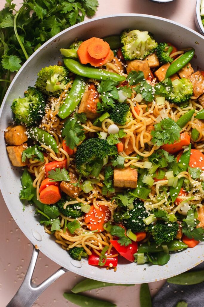 Stir-fried noodles with tofu, broccoli, carrots, and sesame seeds in a pan. Vegan Asian-inspired dish.