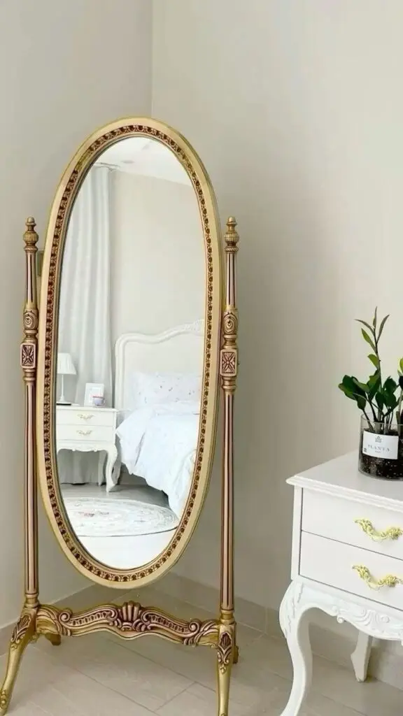 Vintage mirror 🪞 Elegant bedroom mirror with ornate gold frame beside a white nightstand and plant. | Sky Rye Design Elegant bedroom mirror with ornate gold frame beside a white nightstand and plant.