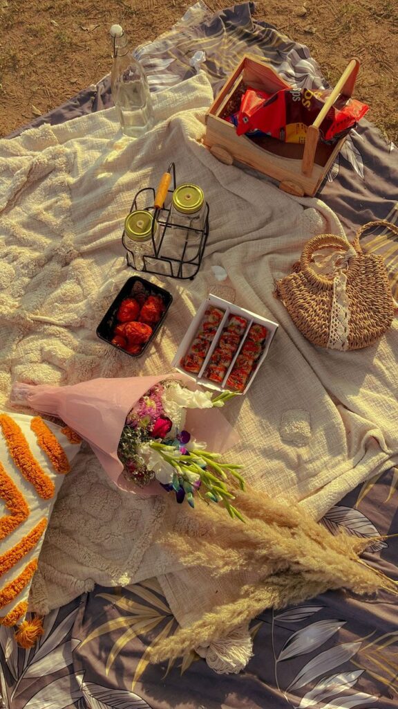 Picnic setup with snacks, sushi, flowers, and a basket on a blanket in sunlight.