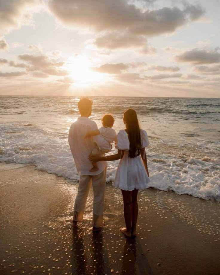 Family enjoying a beach sunset, standing together on the shore in white outfits, facing the ocean waves and golden sky.