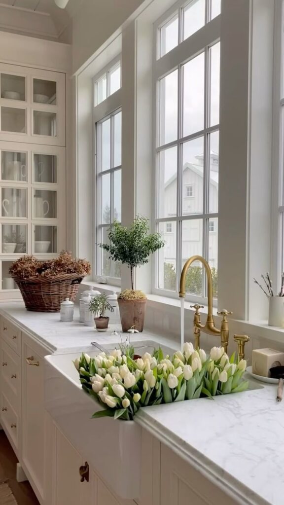 Bright kitchen with large windows, marble counter, gold fixtures, and white tulips in a farmhouse sink.