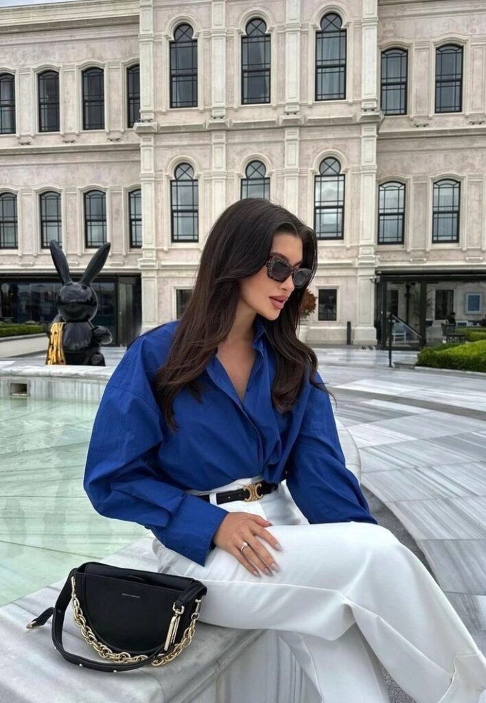 Stylish woman in blue blouse and white pants sitting outdoors near modern building, with designer handbag.