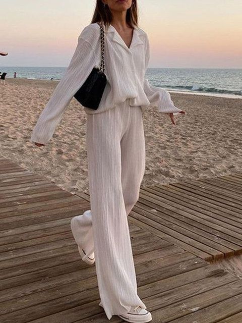 Woman in white outfit walking on beach boardwalk at sunset, carrying black purse.