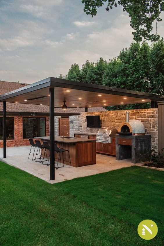 Modern outdoor kitchen with covered patio, grill, bar seating, pizza oven, and lush greenery in a backyard setting.