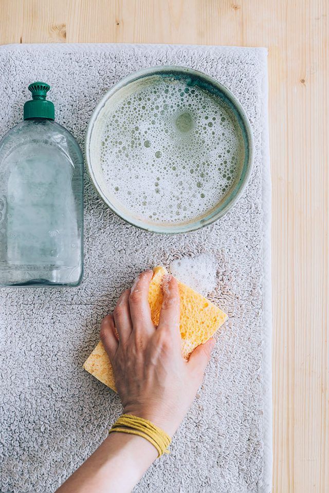 Hand cleaning a towel with soap, a bowl of suds, and a sponge on wooden surface.