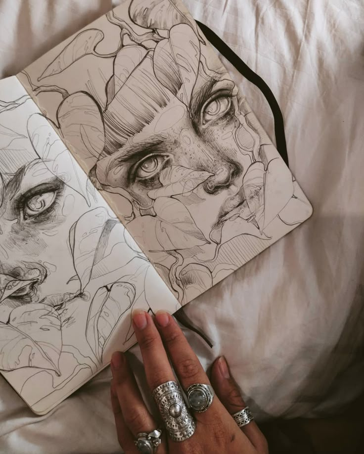 Intricate sketchbook art of a woman's face surrounded by leaves, hand with rings on the page, artistic expression.