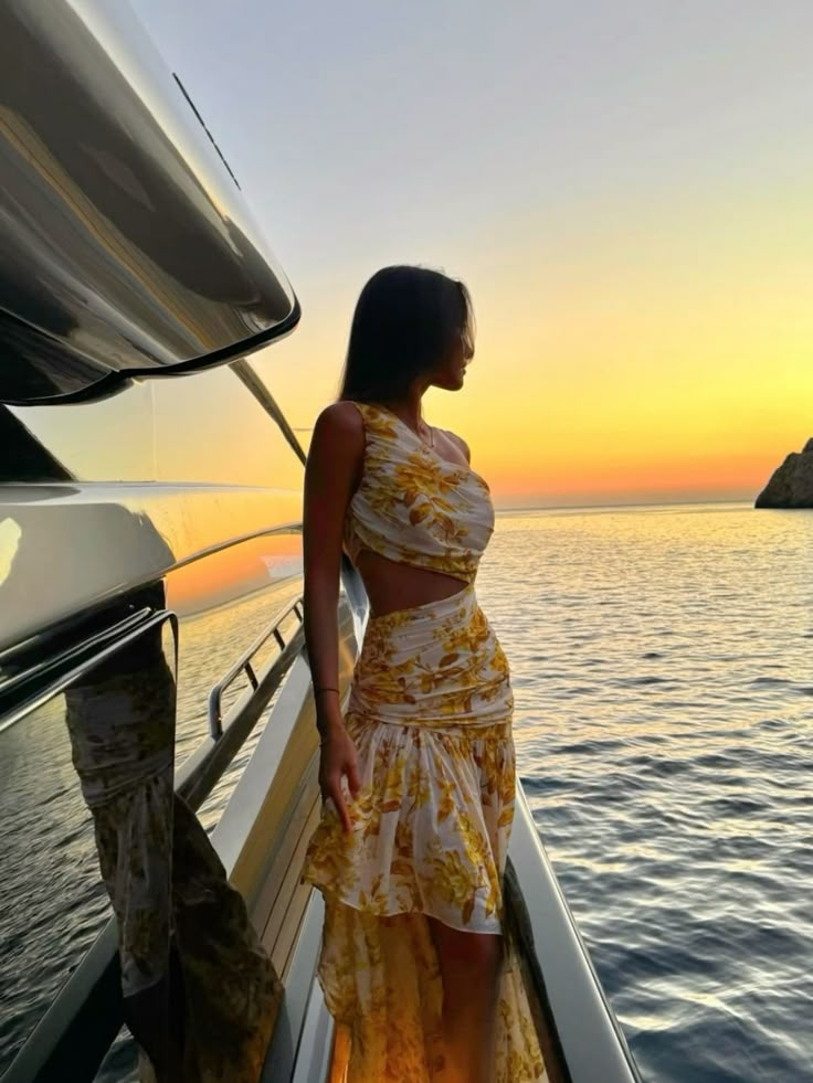 Woman on yacht at sunset, wearing floral dress, gazes at the ocean. Elegant seaside evening scene.