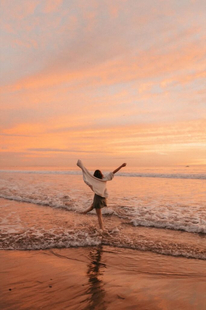 Person joyfully running on beach at sunset with pink sky and ocean waves in the background.