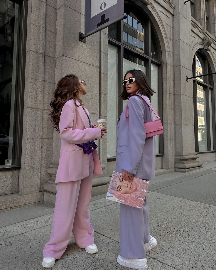 Two stylish women in pastel suits on a city street, holding coffee and a magazine, showcasing trendy fashion.