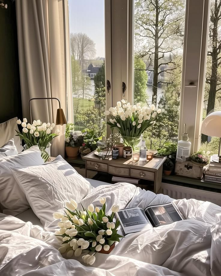 Sunny bedroom with white tulips, cozy bed, and scenic window view of trees and river. Perfect for relaxation.