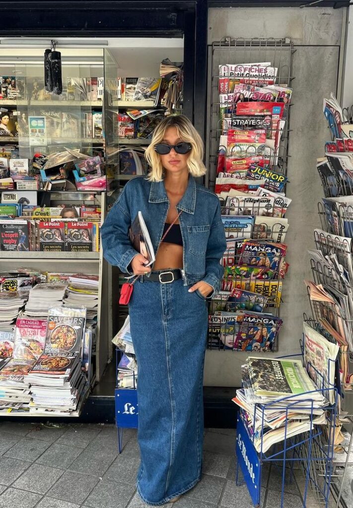 Woman in denim outfit stands by a newsstand, wearing sunglasses and holding a magazine. Stylish urban fashion.