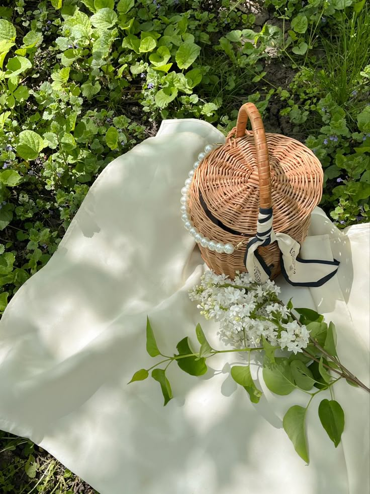 Wicker basket with pearls on a white blanket and flowers, set on green grass in sunlight.