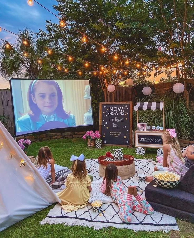 Outdoor movie night with kids watching The Parent Trap on a projector screen, surrounded by cozy blankets and snacks.