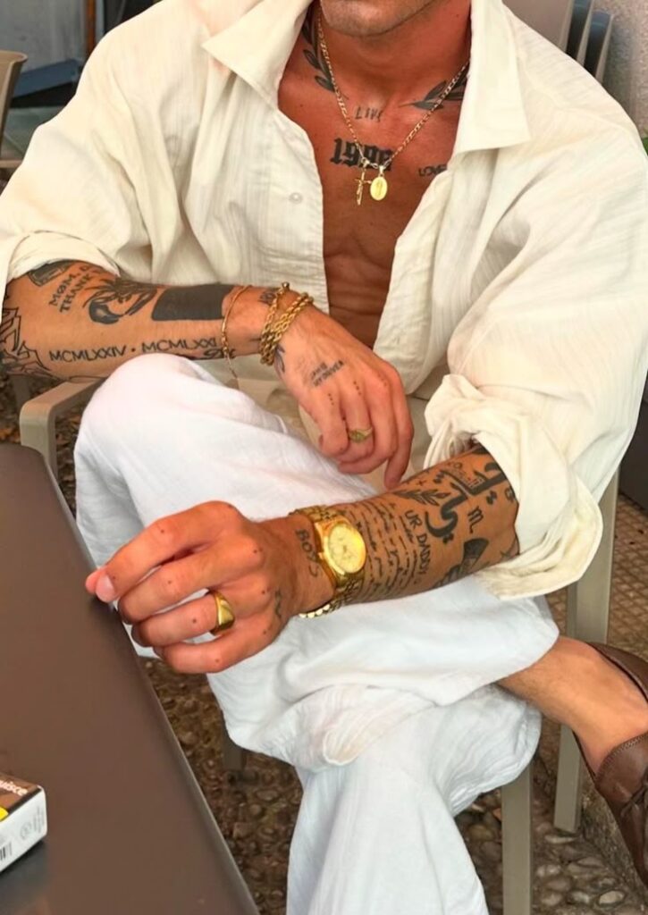 Man in white shirt with tattoos, wearing gold watch and jewelry, sitting casually at a table.