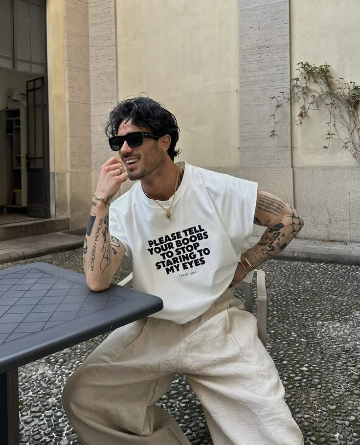 Man with tattoos wearing a humorous T-shirt and sunglasses, sitting at an outdoor table, smiling.