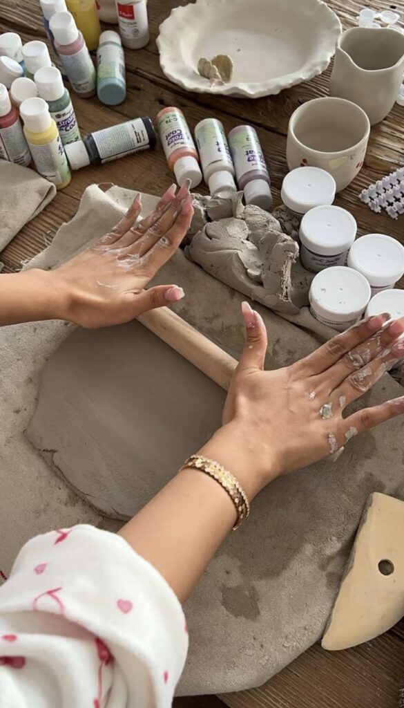 Hands rolling clay on a wooden table with paint bottles, crafts supplies, and pottery tools nearby.