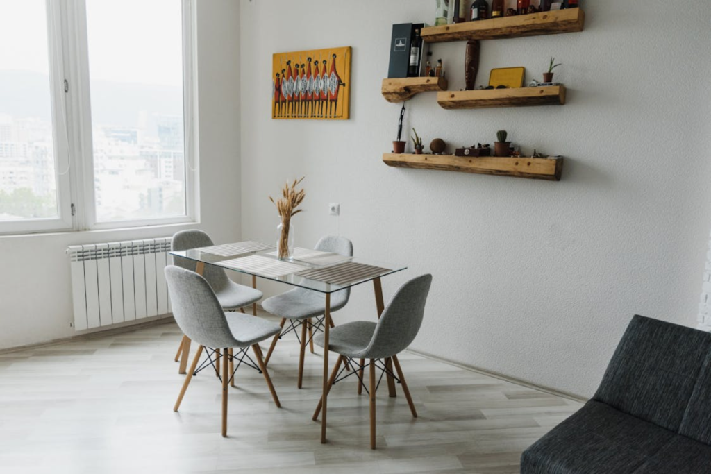 Modern dining room with glass table, gray chairs, wall shelves, and large window overlooking a cityscape.