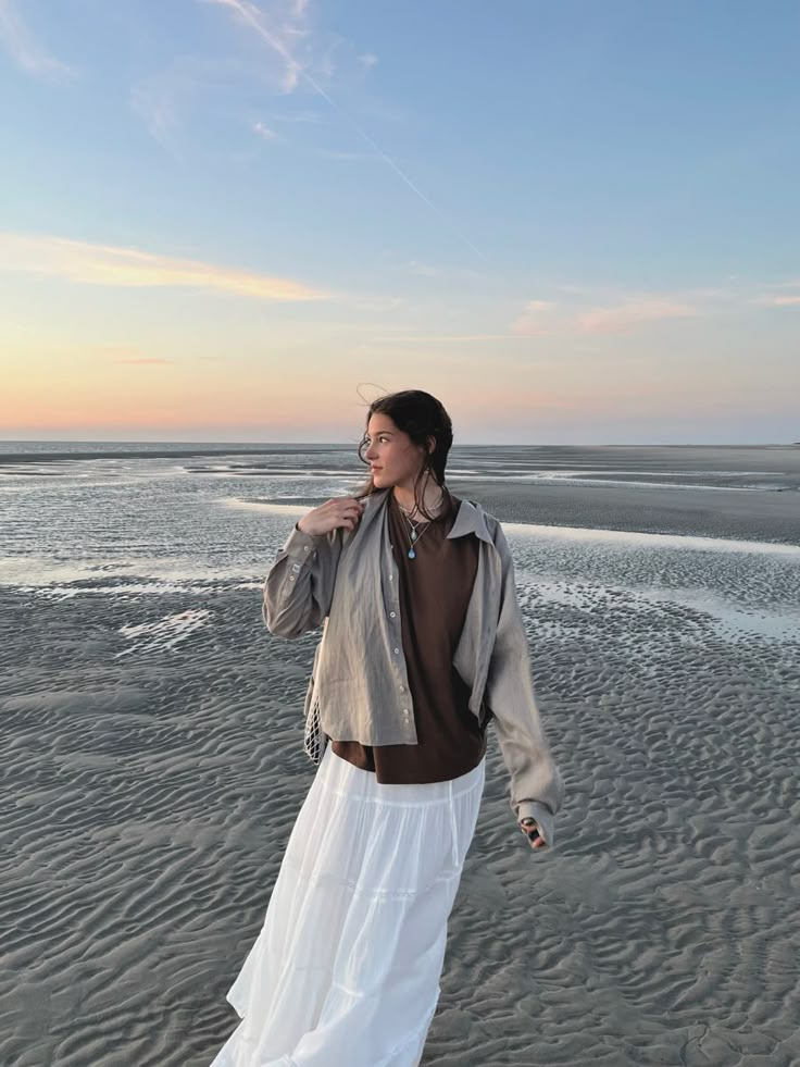 Woman in stylish outfit walking on serene beach at sunset, enjoying coastal scenery.