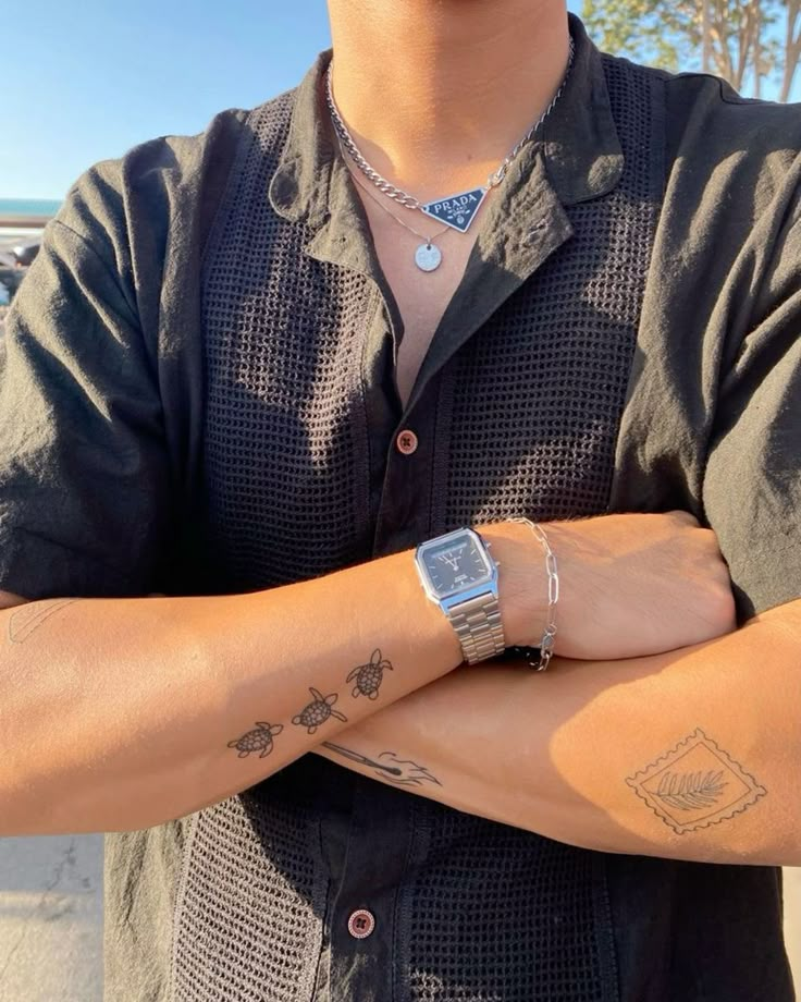 Person in black shirt with silver watch, tattoos, and layered necklaces, standing outdoors.