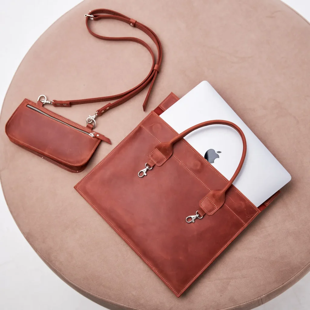 Brown leather purse and matching laptop sleeve on a beige round table, showcasing elegant design and craftsmanship.