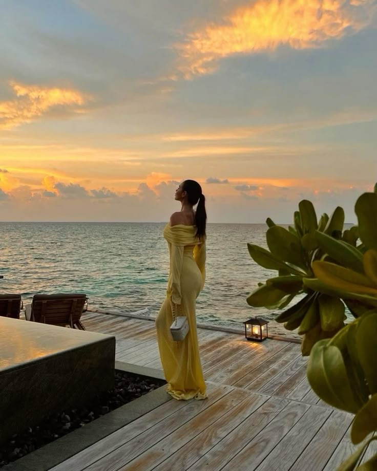 Person in yellow dress on seaside deck at sunset, admiring ocean view and colorful sky.