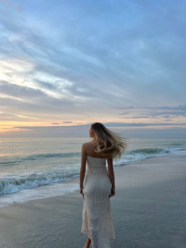 A woman in a white dress walks along the beach at sunset, with waves gently lapping the shore under a colorful sky.