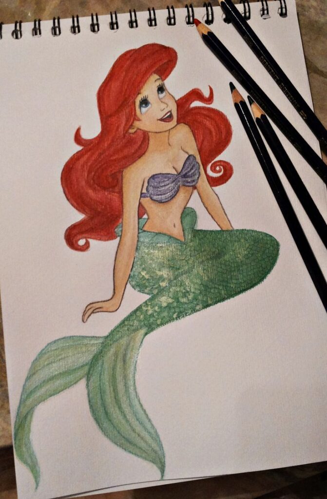 Illustration of a mermaid with red hair, green tail, and purple seashell top, drawn on sketch paper with colored pencils.