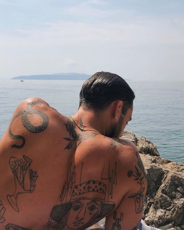 Man with back tattoos sits on rocky shore by the sea, gazing at the water under a clear sky.