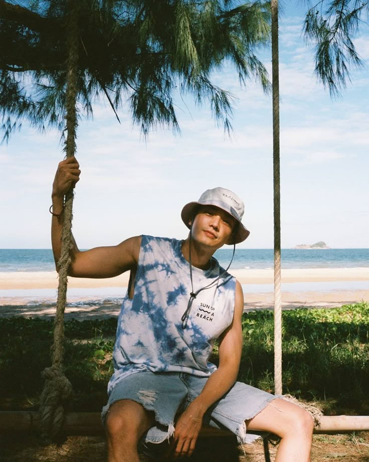 Man relaxing on a beach swing wearing a bucket hat and tie-dye shirt, with ocean and blue sky in the background.