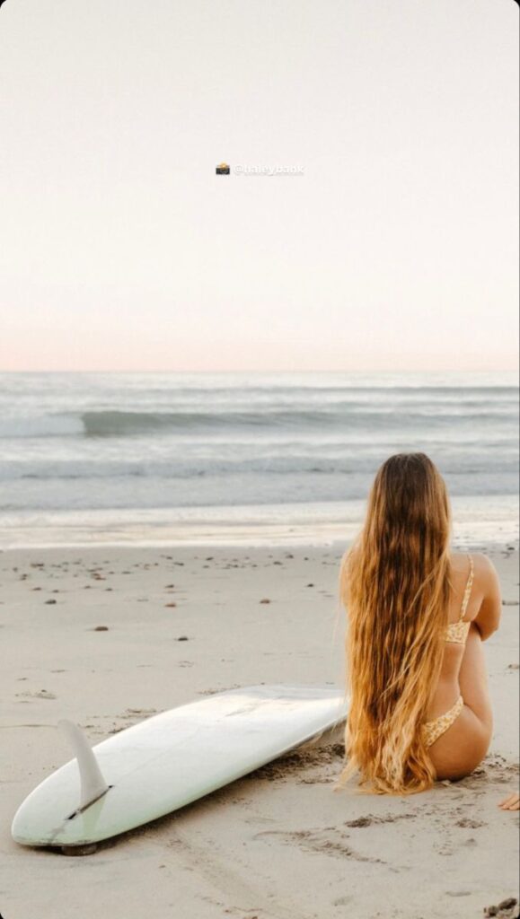 Woman with long hair sitting by the beach with a surfboard, ocean waves in the background.