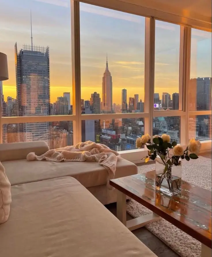 Modern living room with city skyline view at sunset through large windows, featuring a cozy couch and flowers on a table.