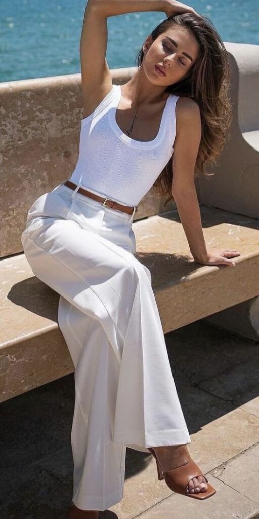 Woman in white summer outfit relaxing by the sea, wearing high-waisted pants and tank top, stylish and elegant look.